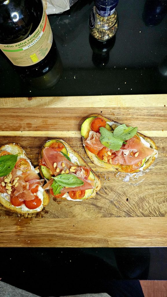 Drie plakjes brood met een combinatie van tomaat, ham, pijnboompitten en basilicum, geserveerd op een houten snijplank.